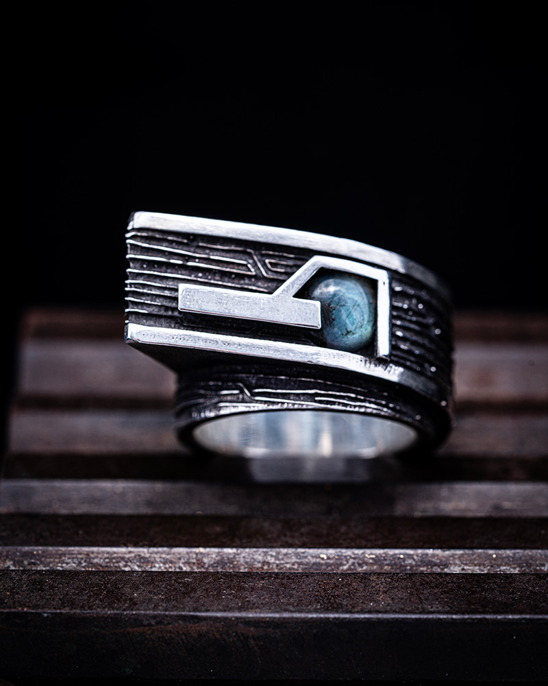 Gravity Ring resting on rusted steel, showing the full width of the engraved sterling silver band and labradorite stone.