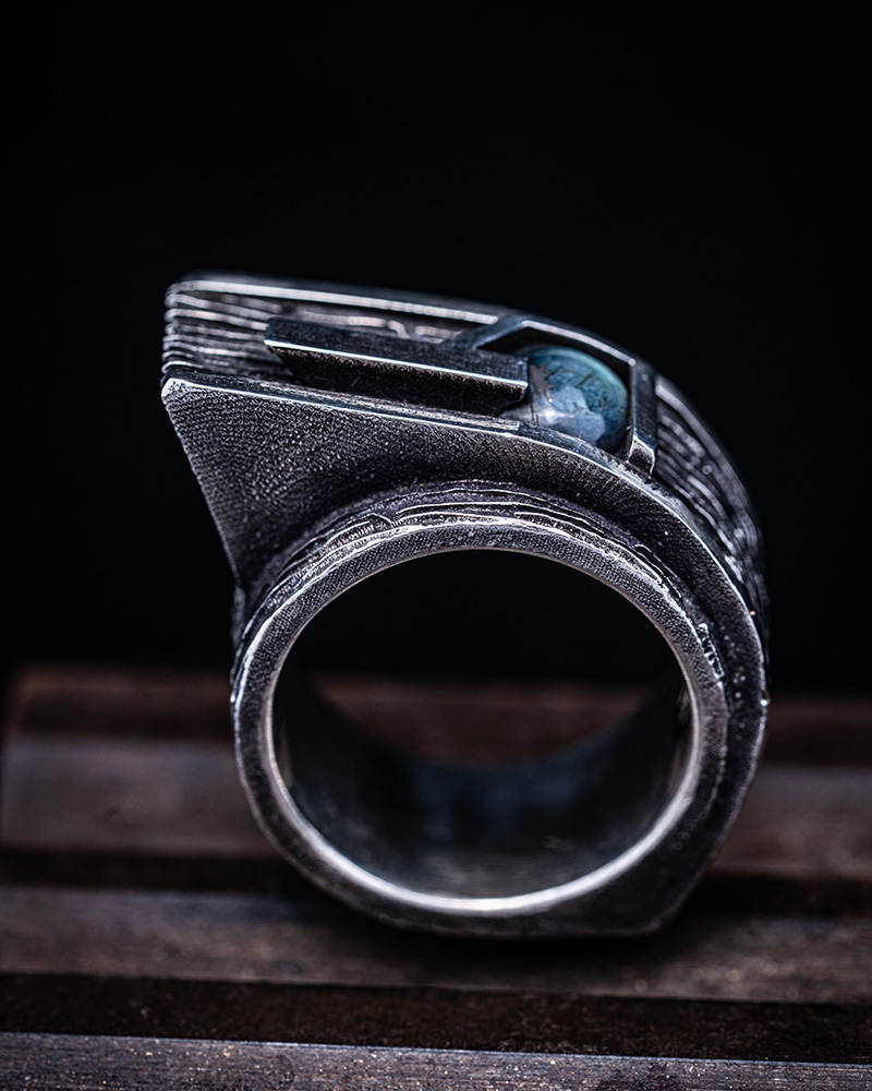 Gravity Ring standing upright on rusted steel, its labradorite cabochon catching the light against the brutalist silver form.