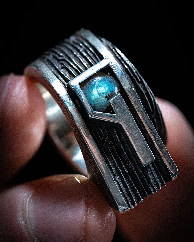 Gravity Ring held between fingertips, showing the engraved linework across the sterling silver band and labradorite cabochon.