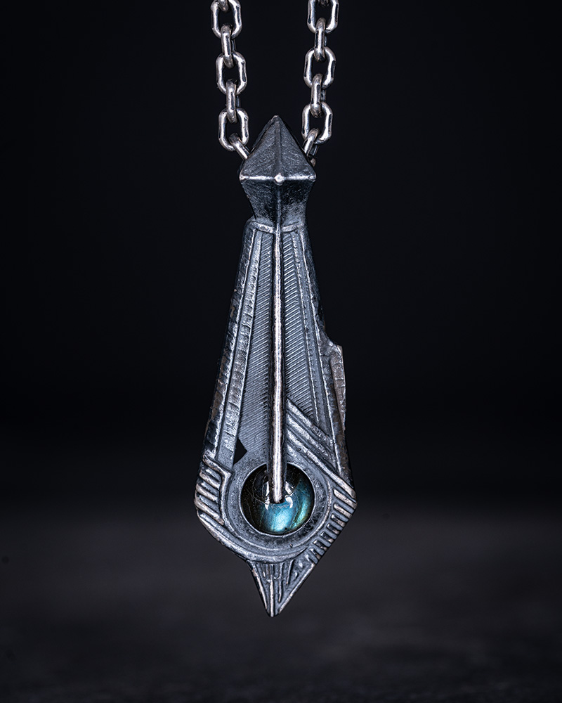 The Memory Pendant against dark slate, the labradorite flashing blue-green at the base of the engraved silver spearhead.