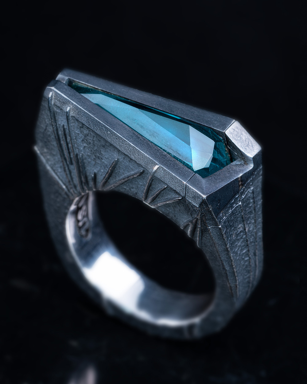The Momentum Ring standing on black stone, its Paraiba tourmaline glowing against the dark oxidized silver.