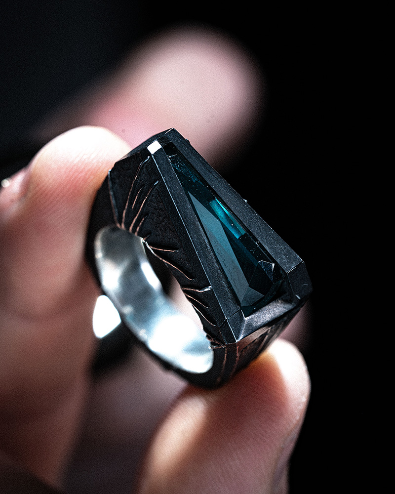 The Momentum Ring held in the fingers in low light, the tapered Paraiba tourmaline catching a blue highlight along its length.