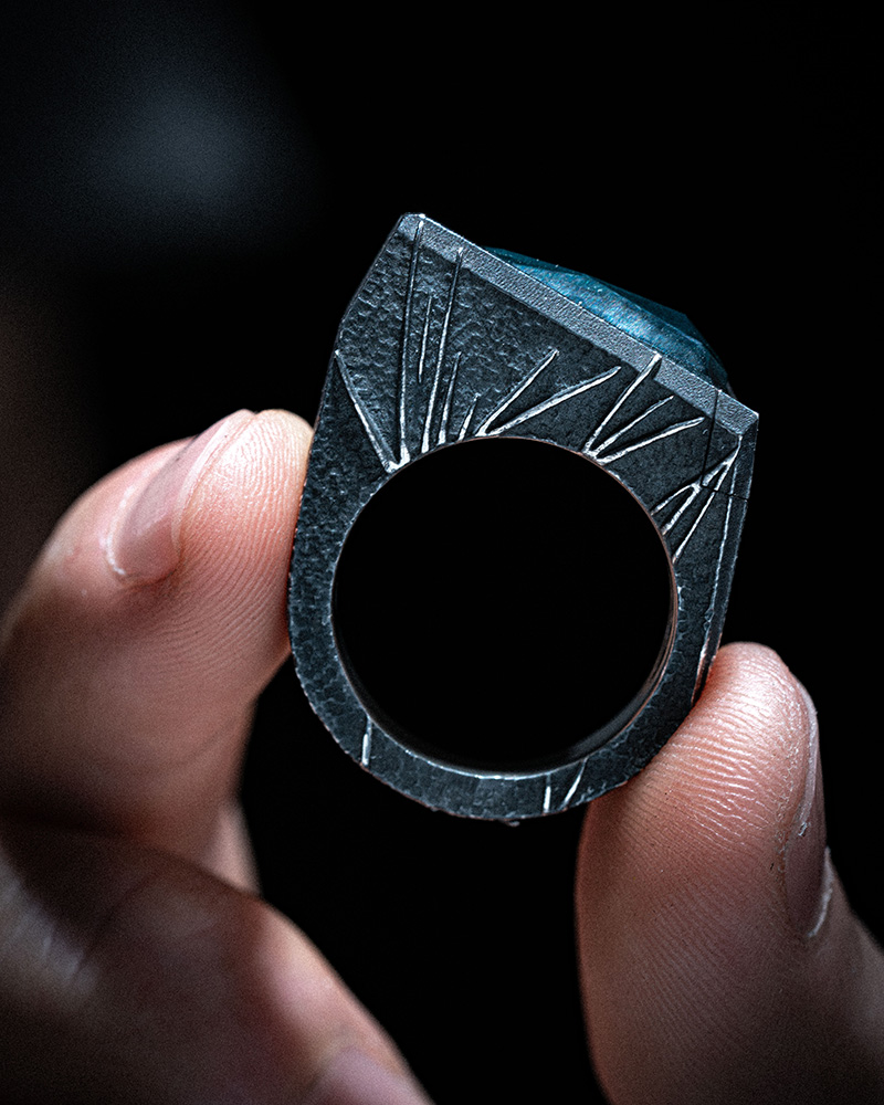 The Momentum Ring held between fingertips from behind, showing the engraved rays fanning out around the band opening.