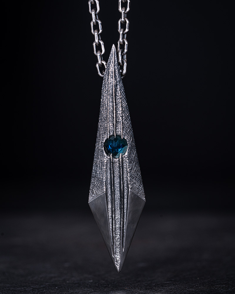 The Shard Pendant photographed against dark slate, its cross-hatched silver facets and Paraiba tourmaline lit from the side.